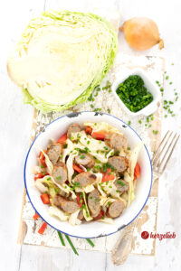 Dieses Bild zum Bratwurstsalat Rezept von herzelieb ist von oben fotografiert. Man schaut in ein weiße Emailleschüssel mit blauem Rand und sieht den Salat aus Spitzkohl, Bratwurstscheiben und gerösteten, roten Paprika. Im Hintergrund eine Schälchen mit Schnittlauchröllchen, ein halbierter Spitzkohl und eine Zwiebel. Rechts vom Teller eine Silbergabel. Im Vordergrund liegen Schnittlauchstängel.