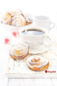 Dieses Foto zum Quarkschnecken Rezept zeigt 2 Quarkschnecken im Vordergrund. Im Hintergrund eine Schale mit mehreren Quarkteilchen, ein Milchkännchen und 2 rosafarbene Blüten