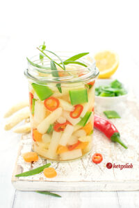 Dieses Foto zum Spargelsalat Rezept zeigt ein Glas mit dem Spargelsalat mit Zuckerschoten, Chiliringen, Karotten und Estragon Im Hintergrund eine rote Chilischote, eine Schale mit Zuckererbse, ein Zitrone und 3 Spargelstangen.