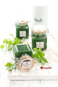 Dieses Foto zum Maggikrautpaste Rezept Zeit drei Gläser mit der Liebstöckelpaste. Eines davon liegt auf der Seite, dahinter stehen zwei weitere Gläser mit der Würzpaste. Ganz im Hintergrund ein Salztopf. Als Dekoration liegen einige Liebstöckelblätter zwischen den Gläsern.