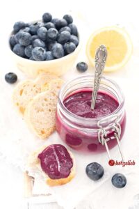 Auf diesem Foto zum Blaubeercreme Aufstrich Rezept sieht man den Heidelbeeraufstrich in einem Weckglas. Vor dem Glas liegt ein bestrichenes Brot. Hinter dem Glas mit dem Brotaufstrich 3 Scheiben Brot, eine gelbe Schale Blaubeeren und eine halbe Zitrone. Dekoriert mit Blaubeeren.