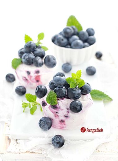 Auf diesem Bild zum Eismuffins Rezept sieht man 2 kleine Eistörtchen Desserts, belegt mit frischen Heidelbeeren und dekoriert mit Zitronenmelisse. Im Hintergrund eine weiße Schale mit Blaubeeren