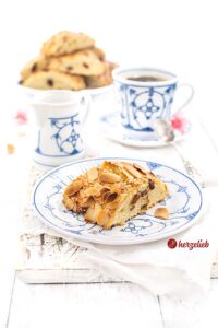 Auf diesem Bild zum dänischen Galopkringle Rezept von herzelieb sieht man ein Stück goldbraun gebackenen Kuchen auf einem blauweißem Teller. Die Kruste ist bestreut mt Zucker und Mandeln. Im Inneren sieht man den hellen Teig mit Rosinen Im Hintergrund ein Milchkännchen, eine Tasse Kaffee und ganz hinten ein Teller mit Kuchenstücken.