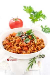 Auf diesem Bild zum Hack-Reis-Topf Rezept sieht man die Hackfleisch-Reispfanne in einer weißen Schüssel. Dekoriert mit blühendem Thymian, einem Zweig Rosmarin und Petersilie. Im Vordergrund liegen Kräuter. Im Hintergrund eine Tomate und ein Glas mit glatter Petersilie
