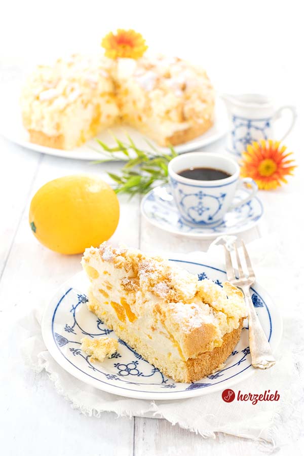 Auf diesem Bild zum Mandarinentorte Rezept sieht man ein Stück Torte auf einem blauweißem Teller. Im Hintergrund eine Tasse Kaffee, eine Orange, der Rest der Torte, ein Milchkännchen und ein orangefarbene Blume