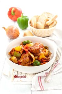 Auf diesem Bild zum Pfannenschlik Rezept von herzelieb sieht man einen weißen, tiefen Teller mit der Schaschikpfanne aus Schnitzelfleisch, Paprika in rot, grün und gelb und Zwiebelspalten. Neben dem Teller liegt eine Silbergabel. Im Hintergrund rechts ein Korb Brot. Links eine Zwiebel, eine rote und grüne Paprikaschote.
