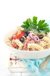 Der Pastalat auf diesem Bild zum Tzatziki Nudelsalat Rezept ist in einer weißen Schüssel angerichtet und dekoriert mit glatter Petersilie, Olivenscheiben, einer Olive, Tomaten und roten Zwiebelringen. Die Schüssel steht auf einem türkisfarbenen Tuch, das auf einem weißen Holzbrett liegt.