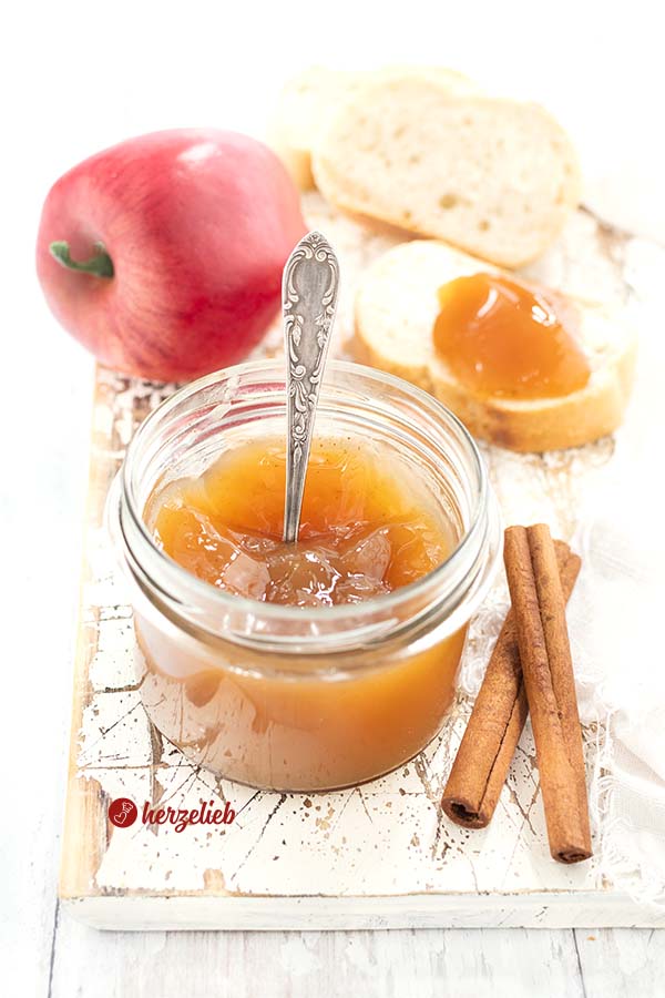 Auf diesem Bild zum Apfelgelee Rezept vom Foodblog herzelieb sieht man ein Glas mit dem Apfelgelee, in dem ein Löffel steck. Im Vordergrund 2 Zimtstangen. Im Hintergrund eine Scheibe Brot mit der Apfelmarmelade, ein Apfel und zwei weitere Scheiben Brort