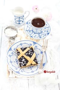 Dieses Foto zum Friesen Pai Rezept ist von oben fotografiert. Man sieht man ein Stück Pflaumenkuchen oder Sylter Friesenpai vom Blech auf einem Teller. Der Kuchen hat eine Mürbeteigboden, eine Füllung aus Pflaumenmus und ein Gitter aus Mürbeteig oben auf. Im Hintergrund eine Tasse Kaffe, ein Milchkännchen und ein silbernes Schälchen mit Hagelzucker. Auf dem Tellerrand liegt eine Gabel.