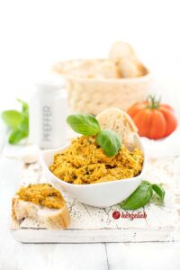 Auf diesem Parmesan-Basilikum Dip sieht an den Käsedip in einer kleinen, weißen Schale. Dekoriert mit Basilikum. Vor dem Schüsselchen liegt ein Stück Brot, dass mit dem Dip bestrichen ist. Im Hintergrund ein Pfefferstreuer, Kräuter, ein Korb Brot und eine Tomate