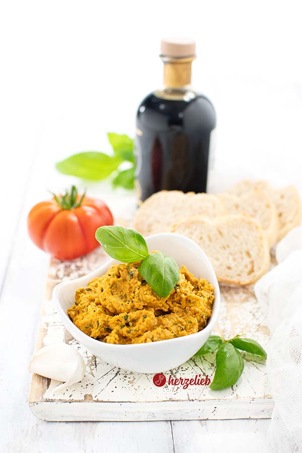Auf diesem Foto zum Parmesan-Basilikum Dip seiht man im Vordergrund ein Schälchen mit dem Käsedip, dekoriert mit Basilikum. Im Hintergrund Brot, eine Flasche dunkler Balsamico, Kräuter und eine Tomate