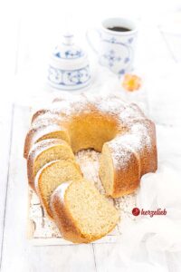 Auf diesem Bild zum Sahne-Gugelhupf Rezept sieht man den angeschnittenen Kuchen auf einem weißen Holzbrett. 3 Scheiben liegen vor den Napfkuchen. Er ist goldgelb gebacken und mit Puderzucker bestäubt. Im Hintergrund ein Zuckertopf und ein Becher Kaffee sowie ein Blüte