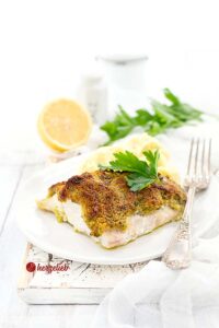 Auf diesem Bild zum Schlemmerfilet Bordelaise Rezept sieht man das Fischfilet mit der Knusperkruste auf einem weißen Teller. Es ist dekoriert mit Petersilie und auf dem Tellerrand liegt eine Silbergabel. Im Hintergrund eine halbe Zitrone, Petersilie, ein Salzstreuer und ein weißes Gewürzgefäß