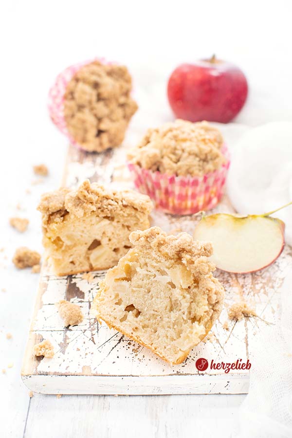 Einen halbierten Muffin sieht man auf diesem Bild zum Apfel-Quarkmuffin Rezept von herzelieb. Rechts dahinter ein Apfelviertel und zwei weitere kleine Apfelkuchen in rot-weißen Muffinförmchen. Ganz hinten ein ganzer, roter Apfel