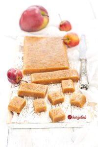 Ein Platte selbstgemachtes Karamell sieht man auf diesem Bild zum Apfelkaramell Rezept. Die Platte ist in Streifen und zu Karamellbonbons geschnitten. Dekoriert in alles mit roten Miniäpfeln, neben der Platte sieht man ein Messer.