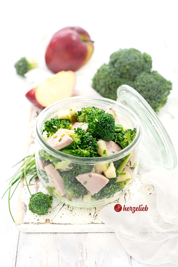 Auf diesem Bild zum Brokkoli-Wurstsalat Rezept sieht man ein Weckglas mit dem Brokkolisalat. An dem Glas lehnt der Glasdeckel. Im Vordergrund ein Brokkoliröschen. Dahinter eine Halme Schnittlauch. Im hitnergrund ein ganzer und ein Viertel Apfel und ein Brokkolikopf