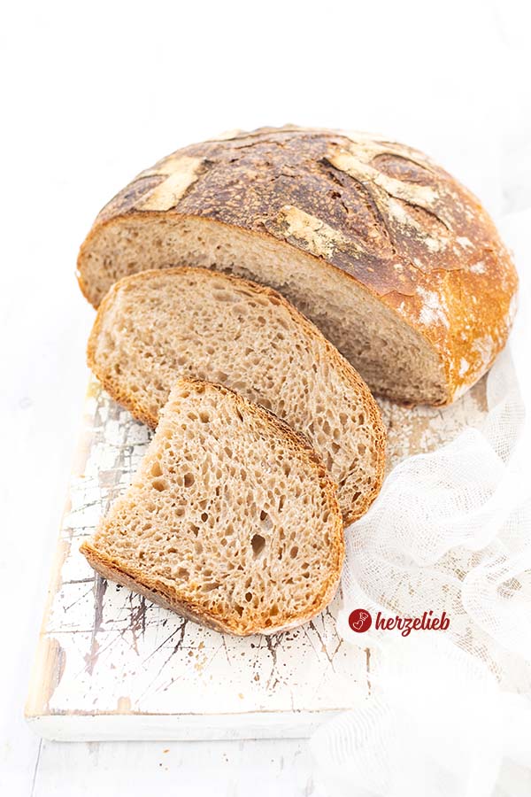 Auf diesem Brot zum Mogelbrot Rezept sieht an ein angeschnittenes Weizenbrot mit knuspriger Kruste und weicher Krume, das auf einem weißen Brett liegt. Der Brotlaib ist angeschnitten und davor liegt eine ganze Scheibe Brot und ein halbe davor 