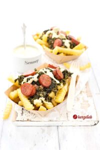 Auf diesem Bild zum Grünkohl to go Rezept sieht man 2 Schalen mit dem Grünkohlgericht. In einer Pappschale sieht man Pommes, Grünkohl, Kohlwurst und Senf-Hollandaise. Auf dem Schalenrand liegt eine Holzgabel. Im Hintergrund, zwischen den Schalen steht ein Schälchen mit dem Dip.