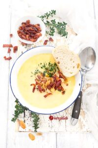 Eine weiße Schüssel mit blauem Rand, gefüllt mit der Käsesuppe sieht man auf diesem Bild zum Käsefondue Suppe Rezept mit einer Prise Liebe von herzelieb. Die Suppe ist dekoriert mit gerösteten Mandeln, Speckwürfeln und einem Thymianzweig. Auf dem Tellerrand liegt eine Scheibe Brot. Rechts neben der Schüssel liegt ein Löffel mit schwarzem Griff, im Vordergrund ein Zweig Thymanzweig. Hinter der Suppenschale steht eine weiße Schale mit Speckwürfelchen, daneben liegt ein Kräuterzweig.