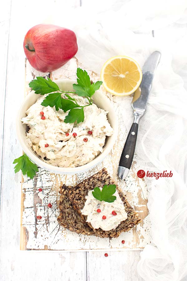 In der Mitte eine Schale mit Brotaufstrich aus Räuchermakrele nach einem Makrelencreme Rezept von herzelieb; rechts ein Messer und eine halbe Zitrone; links hinten ein roter Apfel; vorne rechts zwei Schwarzbrotscheiben, die obere mit Aufstrich bestrichen. Aufstrich und Brot mit Petersilie und roten Pfefferbeeren dekoriert.