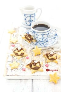 3 Nougatsterne vor einer Tasse und eine Milchkännchen sieht man auf diesem Bild zum Nougatsterne Rezept mit einer Prise Liebe. Die Mürbeteigplätzchen sind gefüllt mit Nougatmasse. Die Weihnachtsplätzchen sind dekoriert mit rosa Zuckersternchen und kleinen Mürbeteigsternen.