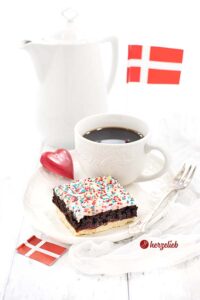 Auf diesem Bild zum Rumkugelschnitten Rezept sieht man ein Stück Kuchen mit einem Mürbeteigboden, einer Schicht Rumkugeln und Streuseln obenauf auf einem weißen Teller. Auf dem Tellerrand eine Kuchengabel. Im Vordergrund ein Dannebrog, im Hintergrund ebenfalls. Hinter dem stück Kuchen steht eine Tasse Kaffee, neben der ein Holzherz liegt. Ganz hinten eine Kaffeekanne.