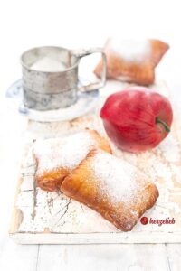 Selbstgemachte, goldbraun gebackene Apfeltaschen auf einem weißen Holzbrett sieht man auf diesem Foto zum Apfelkissen Rezept mit einer Prise Liebe. Dekoriert mit einem roten Apfel, einem silberfarbenen Puderzucker Sieb und einem blauen Kaffeebecher im Hintergrund