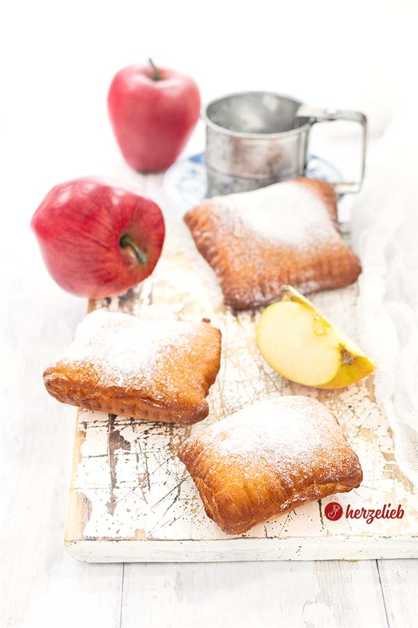 Drei goldbraune Apfeltaschen, bestäubt mit Puderzucker, dekoriert mit einem Apfelviertel und zwei roten Äpfeln im Hintergrund. Daneben liegt ein silberfarbenes Puderzuckersieb.
