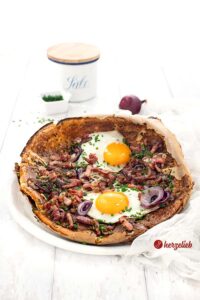 Auf diesem Foto zum herzhaften Ofenpfannkuchen Rezept sieht man Pfannkuchen aus de Backofen auf einem weißen Teller. Belegt mit Speck, roten Zwiebeln und Ei. Im Hintergrund ein Salzfass, eine weiße Schale mit Schnittlauchröllchen und eine rote Zwiebel