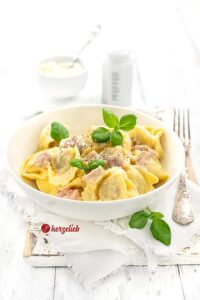 Eine Schale mit Nudeln in Käse-Schinken Soße sieht man auf diesem Bild zum Tortellini alla Panna Rezept mit einer Prise Liebe. Dekoriert mit Basilikumblättern. Rechts neben der Schüssel eine Silbergabel. Hinten ein Pfefferstreuer und eine Schale mit Parmesan