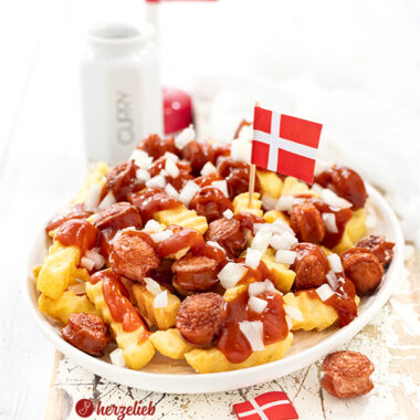 Auf diesem Bild zum Pølsemix Rezept aus Dänemark sieht man das dänische Fastfood Gericht auf einem weißen Teller. In dem Gedicht sieht man Pommes Frites, frittierte Wurstscheiben, Currysauce und rohe Zwiebeln. Im Pølsmix steckt eine dänische Flagge. Im Hintergrund ein weiterer Dannebrog und ein Gewürzgefäß mit Curry