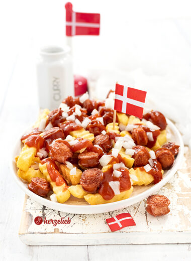 Auf diesem Bild zum Pølsemix Rezept aus Dänemark sieht man das dänische Fastfood Gericht auf einem weißen Teller. In dem Gedicht sieht man Pommes Frites, frittierte Wurstscheiben, Currysauce und rohe Zwiebeln. Im Pølsmix steckt eine dänische Flagge. Im Hintergrund ein weiterer Dannebrog und ein Gewürzgefäß mit Curry