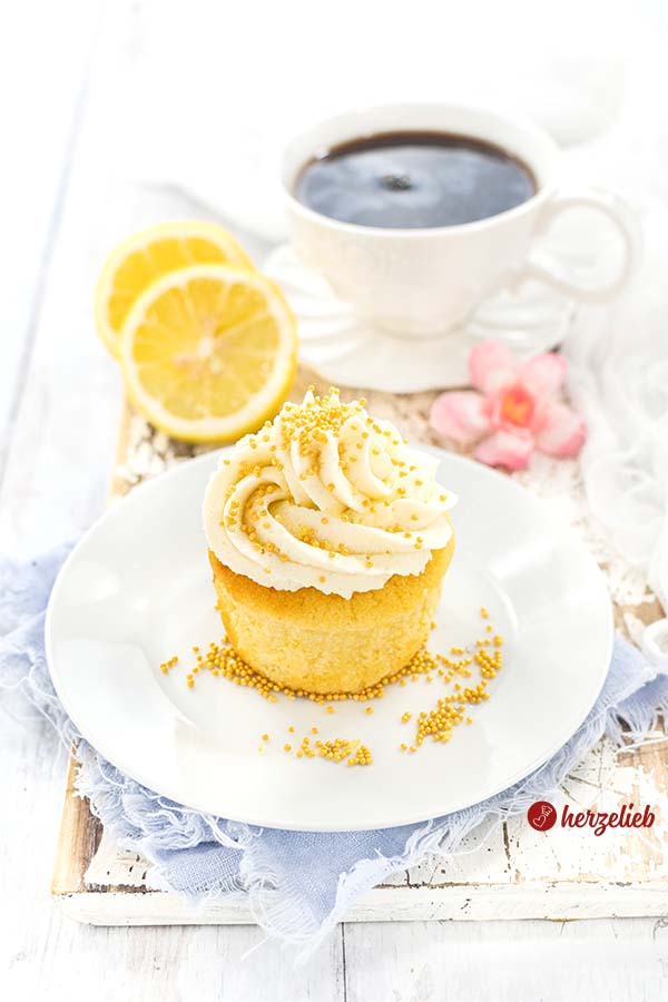 Ein Mini Zitronenkuchen auf einem kleinen, weißen Kuchenteller ist auf diesem Foto zum Zitronen-Cupcakes Rezept. Er ist mit gelben Zuckerperlen bestreut, die auch rundherum auf dem Teller verstreut sind. Im Hintergrund steht eine Tasse Kaffee, daneben liegen 2 Zitronenscheiben.