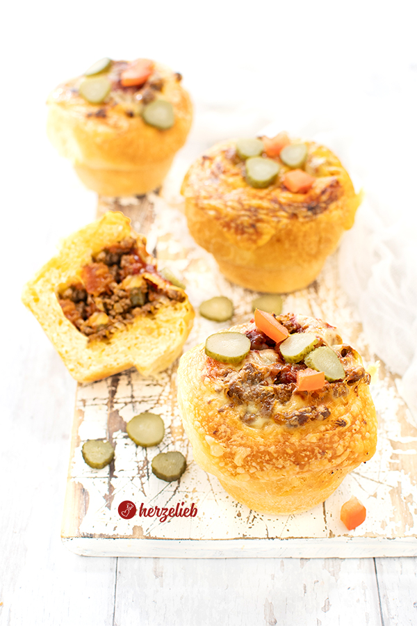 3 ganze und 1/2 herzhafter Muffins sind auf diesem Bild zum Cheeseburger Muffins Rezept zu sehen. Sie sind dekoriert mit Scheiben von Cornichons und Tomatenwürfeln und mit Käse überbacken. Im halben Muffins sieht man die Füllung aus Hackfleisch