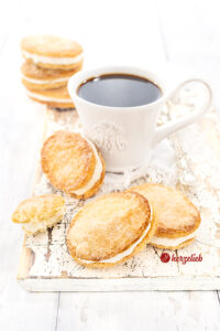 Vorne auf diesem Foto zum Franske Vafler Rezept aus Dänemark sieht man 2 der dänischen Plätzchen. Sie liegen schräg übereinander. Weiter hinten steht eine Tasse Kaffee, an der ein weiterer Keks lehnt. Noch weiter hinten steht ein Stapel aus 3 Franske Vafler.