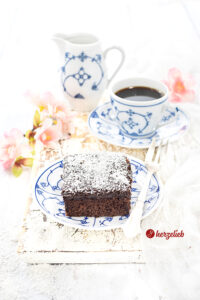 Ein Stück schwedischen Schokoladenkuchen mit Schokoguss sieht man auf diesem Bild zum Kärleksmums Rezept aus Schweden. Im Hintergrund rechts steht eine Tasse Kaffee und ein Milchkännchen. Rechts m Bild liegen Blüten und auf dem Tellerrand liegt eine Kuchengabel aus Silber