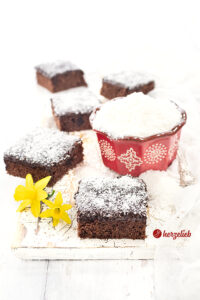 5 Stücke saftigen Schokoladenkuchen aus Schweden sieht man auf diesem Foto zum schwedischen Kärleksmums Rezept. Im Vordergrund liegen zwei Osterglocken Blüten, Zwischen den Schokokuchen Stücken mit dem Schokoladenguss, der mit Kokosflocken bestreut ist, steht eine rote Schale mit Kokosnussflocken, rechts daneben liegt en Silberlöffel