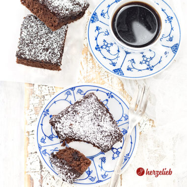 1 Stück schwedischer Schokoladenkuchen auf einem blauweißen Teller sieht man auf diesem Bild zum Kärleksmums Rezept aus Schweden. Im Hintergrund steht steht eine Tasse Kaffee, links davon liegen zwei weitere Stücke Schokoladenkuchen