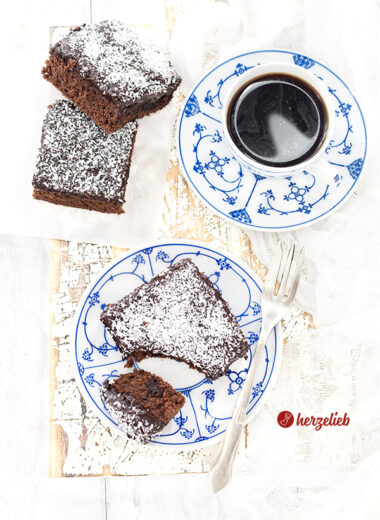1 Stück schwedischer Schokoladenkuchen auf einem blauweißen Teller sieht man auf diesem Bild zum Kärleksmums Rezept aus Schweden. Im Hintergrund steht steht eine Tasse Kaffee, links davon liegen zwei weitere Stücke Schokoladenkuchen