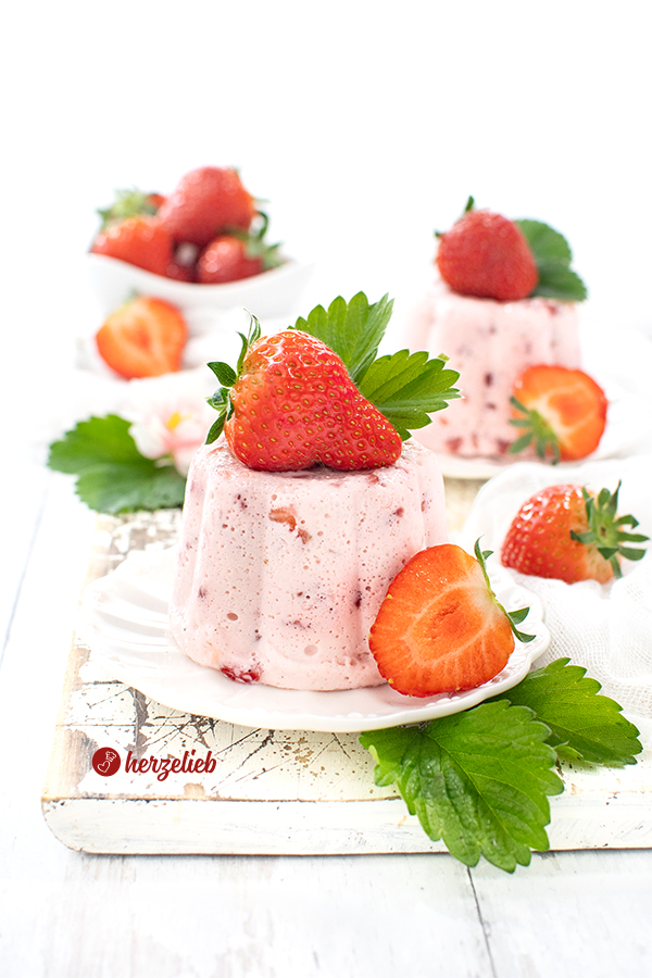 Zwei Erdbeer-Joghurt-Dessert auf einem kleinen weißen Teller sieht man auf diesem Foto zum Erdbeernachtisch Rezept von herzelieb. Beide sind dekoriert mit frischen Erdbeeren und Erdbeerblättern. Weiter hinten sieht man eine kleine weiße Schale mit ganzen Früchten.