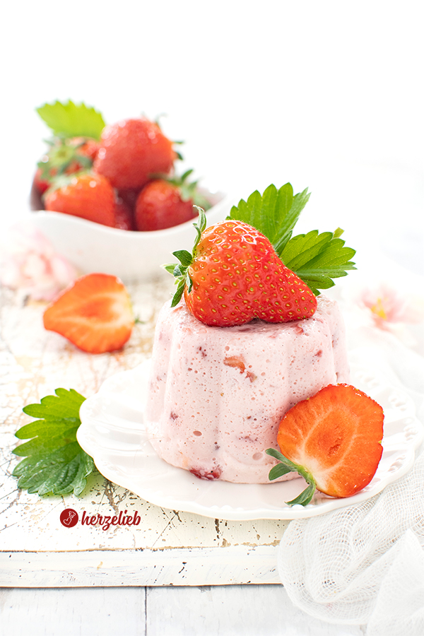 Einen weißen kleinen Teller sieht man auf diesem Foto zum Erdbeer-Joghurt-Dessert Rezept von herzelieb. Man schaut von schräg vorne auf den Erdbeernachtisch. Dekoriert ist es mit einer ganzen Erdbeere und einem Erdbeerblatt. Hintergrund steht eine weiße Schale mit Erdbeeren.