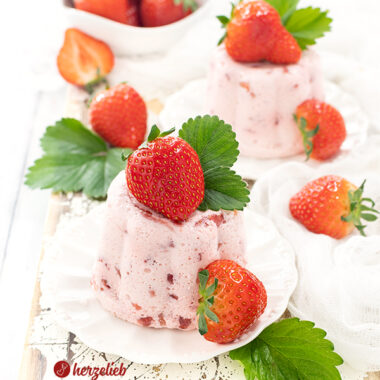 Zwei kleine, weiße Teller mit einem Erdbeer-Nachtisch sieht man auf diesem Bild zum Erdbeer-Joghurt-Dessert Rezept von herzelieb. Sie sind leicht rosa und man sieht darin kleine Erdbeerstückchen. Dekoriert sind beide Desserts mit ganzen Erdbeeren und Erdbeerblättern.