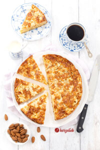 Einn goldbraun gebackener und angeschnittener Kuchen mit Knusperkruse mit Mandeln aus der Springform Springform nach einem Hummellkuchen Rezept von herzelieb. Im Hintergrund eine Tasse Kaffee und ein Kuchenstück. Rechts vom Teller ein Messer und vorne links eine Schale Mandelnö