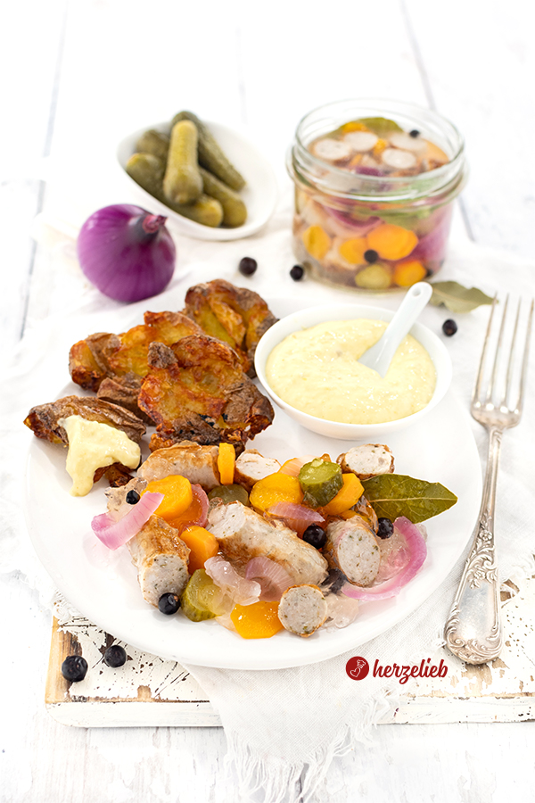 Bratwurstsülze mit Quetschkartoffeln und selbstgemachter Remoulade auf einem ovalen Teller. Rechts vom teller liegt eine Gabel. Im Hintergrund eine rote Zwiebel, ein Schälchen mit Gewürzgurken und ein Glas mit der selbstgemachten Sülze