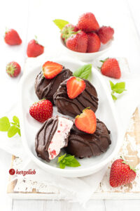 Eiskonfekt nach einem Eispralinen Rezept von herzelieb auf einer weißen länglichen Platte. Als flache Scheiben mit Schokolade überzogen. Dekoriert mit Erdbeeren und Zitronenmelisse.