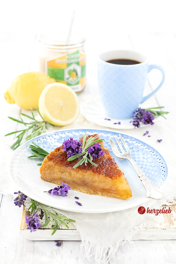 Ein Stück Honigkuchen nach einem Imkerkuchen rezept von herzelieb. Dekoriert mit Lavenblüten und Lavendelblättern. Im Hintergrund eine halben und eine ganze Zitrone, ein Glas Honig und eine Tasse Kaffee