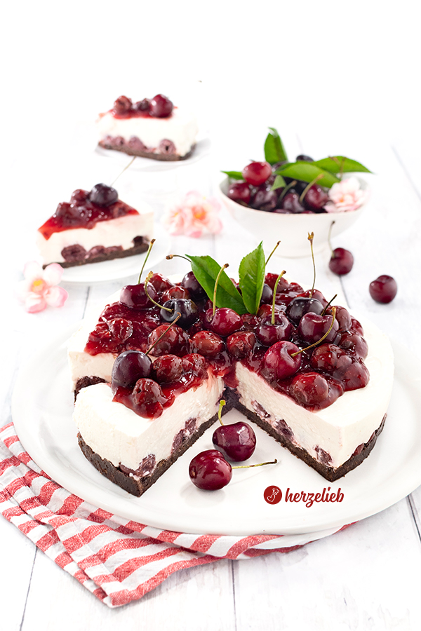 Angeschnittene Schwarzbrottorte mit Quarkcreme und Kirschen nach einem Pumpernickeltorte Rezept von herzelieb. Dekoriert mit frischen Kirchen. Im Hintergrund 2 Teller mit Kuchenstücken und eine Schale Kirschen. Im Vordergrund ein rotweiß gestreiftes Tuch.