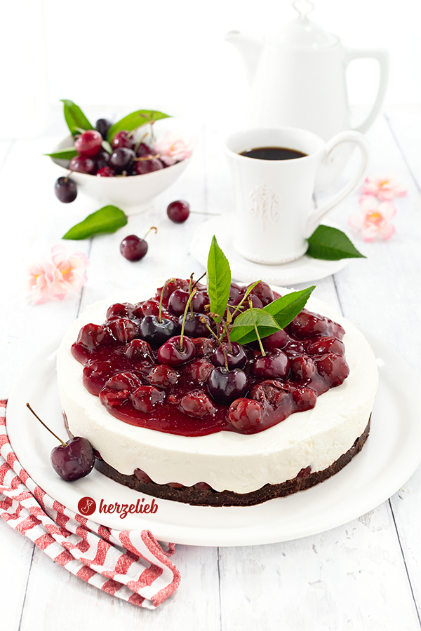 Eine ganze Kühlschranktorte auf einem weißen Tortenteller nach einen Pumpernickeltorte Rezept mit Kirschen und Quarkcreme von herzelieb. Dekoriert mit ganze Kirsche, Blättern vom Kirschbaum. Im Hintergrund eine Schale Früchte und eine Tasse Kaffee.