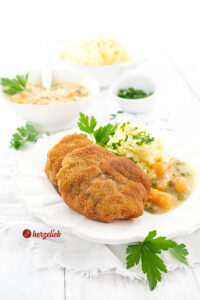 Zwei panierte Hackfleischschnitzel auf einen weißen Teller. Angerichtet mit Kartoffelbrei und Erbsen und Wurzeln in weißer Soße, dekoriert mit Petersilie. Im Hintergrund eine Schüssel mit Kartoffelmus, eine mit Gemüse in weißer Sauce und ein Schälchen mit gehackter Petersilie