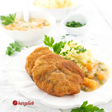 Zwei panierte Hackfleischschnitzel auf einen weißen Teller. Angerichtet mit Kartoffelbrei und Erbsen und Wurzeln in weißer Soße, dekoriert mit Petersilie. Im Hintergrund eine Schüssel mit Kartoffelmus, eine mit Gemüse in weißer Sauce und ein Schälchen mit gehackter Petersilie